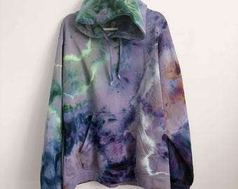 May include: A long-sleeve hooded sweatshirt featuring a tie-dye pattern in shades of lavender, deep purple, forest green, and bright lime. The garment includes a front kangaroo pocket, a drawstring hood, and ribbed cuffs at the sleeves and hem.