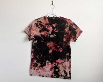 May include: A short-sleeve crew neck t-shirt featuring a tie-dye pattern in shades of black, deep red, and muted pink. The garment is displayed on a white plastic hanger, showcasing a marbled, abstract design across the front and sleeves.