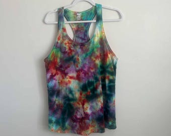 May include: A sleeveless racerback tank top featuring a vibrant tie-dye pattern with blended swirls of red, purple, teal, yellow, and dark green colors. The garment is displayed on a white plastic hanger against a plain, neutral-colored wall.