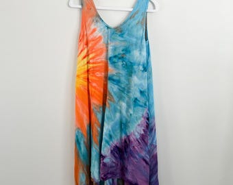 May include: A sleeveless tie-dye dress with a V-neck. The dress features a vibrant color scheme, with orange and yellow on one side and blue and purple on the other. The dress is hanging on a white hanger.