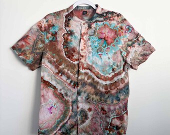 May include: A short-sleeve button-down shirt featuring a geode-inspired tie-dye pattern in shades of brown, pink, teal, and white. The shirt has a mandarin collar, a single chest pocket, and a button-front closure, displaying organic, swirling shapes.