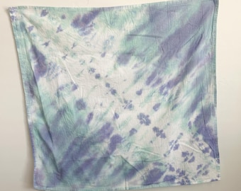 May include: A square fabric cloth featuring a tie-dye pattern in shades of light purple, mint green, and white. The design displays a diagonal orientation with blended color transitions and soft, organic shapes across the entire surface area.