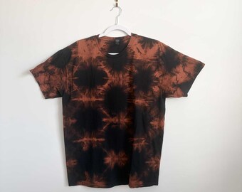 May include: A short-sleeve crew neck t-shirt featuring a black and rust-colored tie-dye pattern. The design consists of repeating circular sunburst motifs across the fabric, creating a high-contrast aesthetic on the front of the garment.
