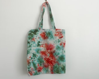 May include: A tie-dye tote bag with a vibrant pattern of red, green, and light blue hues. The bag is hanging against a white wall. The tote bag has matching handles and a rectangular shape, suitable for carrying various items.