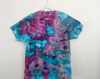 May include: A short-sleeved t-shirt with a tie-dye pattern in shades of teal, purple, and pink. The shirt is hanging on a white hanger against a white wall. The tie-dye design creates a unique and colorful look.