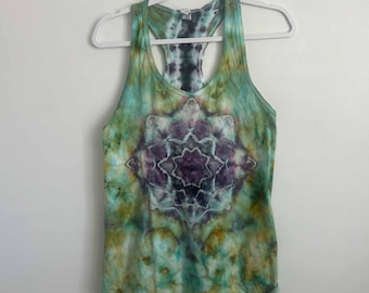 May include: A racerback tank top featuring a tie-dye pattern in shades of teal, olive green, and deep purple. The center of the garment displays a symmetrical, star-shaped mandala design in dark purple and white, set against a marbled background.