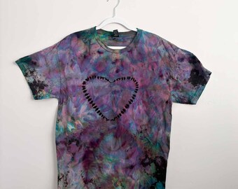 May include: A short-sleeve crew neck t-shirt featuring a tie-dye pattern in shades of purple, blue, and black. The center of the shirt displays a heart shape outlined by a series of small, dark, individual marks against the colorful background.