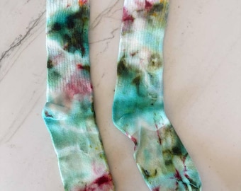 May include: A pair of crew-length socks featuring a tie-dye pattern with blended shades of teal, light green, and magenta. The socks have a ribbed texture along the upper portion and a smooth knit finish throughout the foot and heel area.