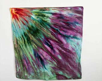 May include: A square silk scarf featuring a vibrant tie-dye pattern that radiates from the top left corner. The design displays a blend of deep magenta, forest green, turquoise, and bright yellow hues, creating a fluid, painterly effect.