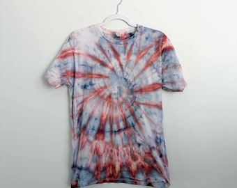 May include: A short-sleeve crew neck t-shirt featuring a spiral tie-dye pattern in shades of red, blue, and white. The garment is displayed on a white plastic hanger against a plain background, showcasing a circular color distribution.