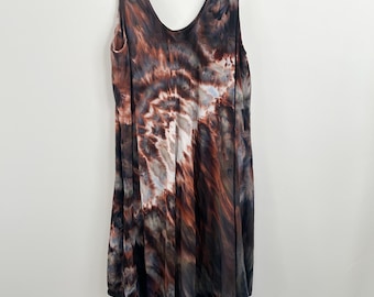 May include: A sleeveless, V-neck dress with a tie-dye pattern in shades of brown, black, and white. The dress hangs from a white hanger against a white background. The fabric appears soft and flowing, with an asymmetrical hemline.