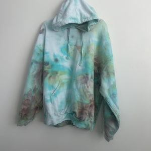 May include: A tie-dye hoodie in shades of blue, green, and brown hangs against a white wall. The hoodie features a drawstring hood and a front pocket. The fabric has a marbled effect, with the colors blending together in a unique pattern. The garment is casual wear.