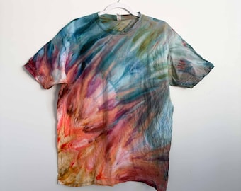 May include: A short-sleeve crew neck t-shirt featuring a multi-colored tie-dye pattern. The design incorporates blended swirls of vibrant orange, deep red, muted teal, and soft blue hues across the fabric, creating a fluid and abstract appearance.
