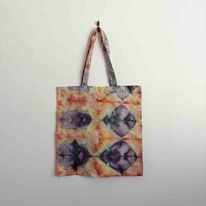 May include: A rectangular fabric tote bag featuring a tie-dye pattern with blended shades of purple, yellow, orange, and red. The design displays repeating diamond-shaped motifs across the surface, complemented by two matching fabric handles.