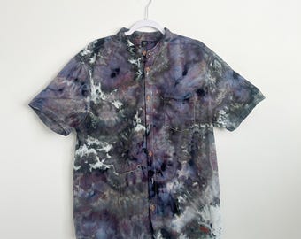 May include: A short-sleeved button-down shirt featuring a dark tie-dye pattern in shades of purple, black, gray, and white. The shirt has a mandarin collar, a single chest pocket on the left side, and a row of small buttons down the center front.