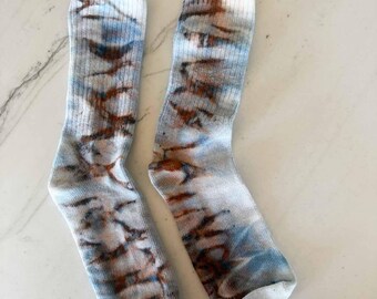 May include: A pair of crew-length socks featuring a ribbed texture and a tie-dye pattern in shades of light blue, white, and earthy brown. The socks have a consistent vertical design with blended colors throughout the fabric from the cuff to the toe.