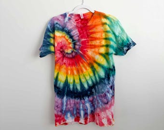 May include: A short-sleeve crew neck t-shirt featuring a vibrant multi-colored tie-dye pattern. The design displays a prominent spiral motif radiating from the center, incorporating bold shades of red, orange, yellow, green, blue, and purple.