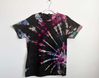 May include: A short-sleeve crew neck t-shirt featuring a black base with a vibrant tie-dye spiral pattern in shades of magenta, bright blue, and white. The design radiates from the lower right side across the front of the garment.