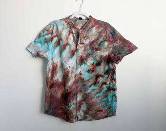 May include: A short-sleeve button-down shirt featuring a multi-colored tie-dye pattern in shades of teal, brown, and muted red. The shirt has a mandarin collar, a single chest pocket on the left side, and a small rectangular label at the bottom hem.