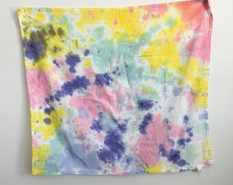 May include: A square fabric cloth featuring a tie-dye pattern with blended colors of bright yellow, soft pink, light blue, and deep purple. The textile has a visible woven texture and a small white fabric tag attached to the bottom right corner.