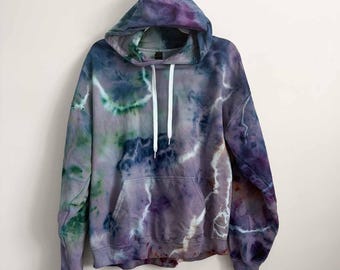 May include: A long-sleeve hooded sweatshirt featuring a tie-dye pattern in shades of purple, blue, and green. The garment includes a front kangaroo pocket, a drawstring hood with white cords, and ribbed cuffs at the sleeves and bottom hem.
