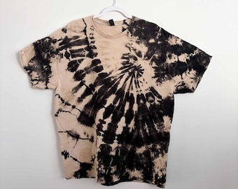 May include: A short-sleeve crew neck t-shirt featuring a black and beige tie-dye spiral pattern. The design radiates from the right side of the chest across the front of the garment, creating a high-contrast swirl effect against the light base.