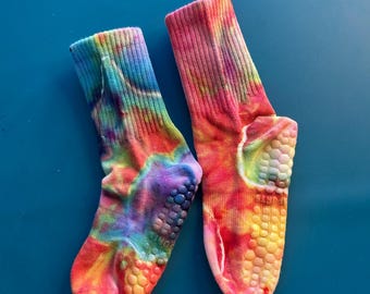 May include: A pair of crew-length socks featuring a vibrant tie-dye pattern in shades of pink, orange, yellow, blue, and purple. The soles of the socks are equipped with a textured, non-slip grip pattern made of small, raised circular silicone dots.