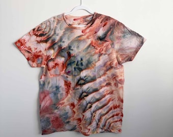 May include: A short-sleeve crew neck t-shirt featuring an abstract tie-dye pattern in shades of muted red, dusty rose, slate blue, and charcoal gray. The garment is displayed on a white plastic hanger against a plain, light-colored wall.