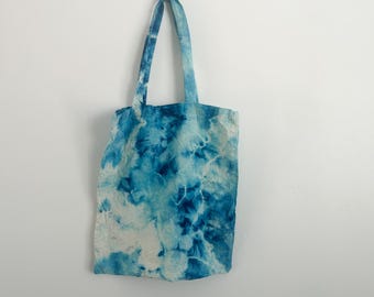 May include: A blue and white tie-dye tote bag hanging against a white wall. The bag has a rectangular shape with two handles and a pattern of swirling blue and white colors. The bag is made of fabric.