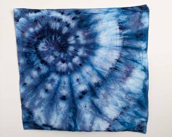 May include: A square scarf featuring a spiral tie-dye pattern in various shades of blue, ranging from deep navy to light sky blue. The fabric displays a radiating design with darker indigo concentrations at the center, fading into lighter white and blue tones toward the edges.