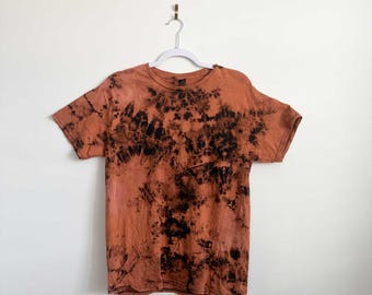 May include: A short-sleeve crew neck t-shirt featuring a tie-dye pattern in shades of burnt orange and black. The garment has a round neckline, short sleeves, and a straight hemline, displaying a mottled, dark, and earthy color combination throughout.