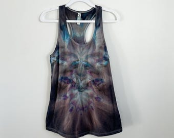 May include: A racerback tank top featuring a tie-dye pattern in shades of dark gray, muted blue, and purple. The garment has a scoop neckline and is displayed on a white plastic hanger against a plain, light-colored wall background.