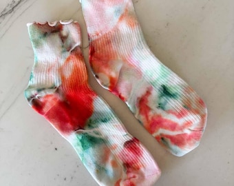 May include: A pair of ribbed ankle socks featuring a tie-dye pattern in vibrant shades of red, orange, green, and white. The socks have a distinct lettuce-edge ruffle trim at the top cuff, providing a textured finish to the overall design.