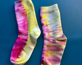 May include: A pair of ankle-length socks featuring a tie-dye pattern in vibrant shades of hot pink and bright yellow. The socks have a ribbed cuff at the top and a soft, blended appearance where the colors transition across the fabric surface.