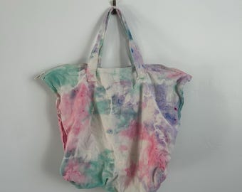 May include: A tie-dye tote bag featuring a soft color palette of pastel pink, teal, and purple swirls on a white base. The bag includes two fabric handles at the top and a horizontal zipper closure along the upper edge for securing contents.