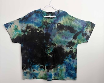 May include: A short-sleeve t-shirt featuring a tie-dye pattern in shades of black, deep blue, teal, and light green. The garment has a round neckline and a loose, boxy fit, with the dark dye concentrated in the center and lighter tones at the edges.