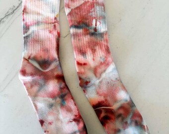 May include: A pair of ribbed crew socks featuring a tie-dye pattern with blended shades of muted red, soft pink, grey, and white. The socks have a vertical ribbed texture throughout the leg and foot, with a marbled, watercolor-like appearance.