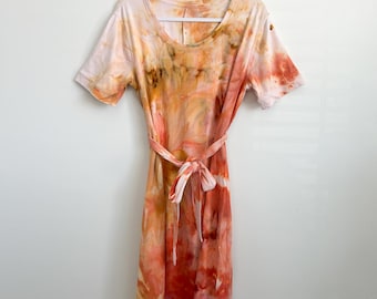 May include: A short-sleeve midi dress featuring a marbled tie-dye pattern in shades of peach, coral, orange, and cream. The dress has a round neckline and a matching fabric tie belt at the waist, hanging on a white plastic hanger.
