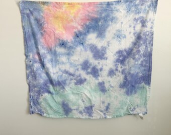 May include: A square fabric cloth featuring a tie-dye pattern with blended colors of pastel pink, soft yellow, light blue, deep violet, and mint green. The textile has a visible woven texture and a crinkled appearance across the surface.