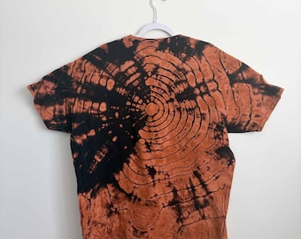 May include: A short-sleeve t-shirt featuring a black and burnt orange tie-dye pattern that resembles the concentric rings of a tree trunk. The shirt is displayed hanging on a white plastic hanger against a plain, light-colored wall background.