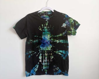 May include: A short-sleeve t-shirt featuring a black base with a central tie-dye pattern in shades of blue, teal, and green. The design radiates outward from the center in a symmetrical, geometric style, creating a stark contrast against the black fabric.