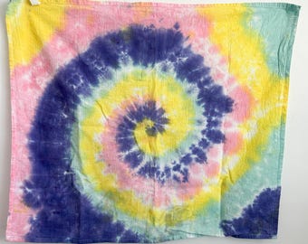 May include: A square fabric cloth featuring a spiral tie-dye pattern in vibrant shades of deep purple, bright yellow, soft pink, and light teal. The design radiates from the center in a circular motion, covering the entire surface of the textile.