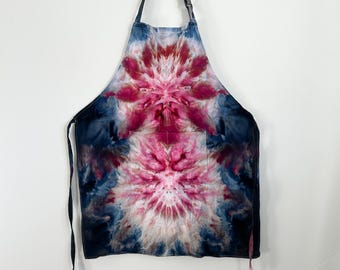 May include: A tie-dye apron featuring a vibrant pattern of deep magenta and pink starburst designs set against a dark navy blue background. The apron includes an adjustable neck strap with a black buckle and a large front pocket.