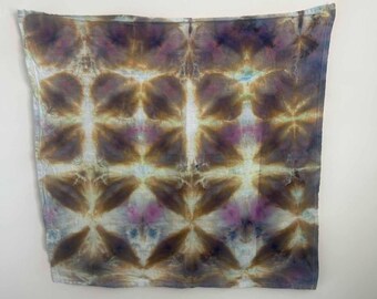 May include: A square fabric cloth featuring a tie-dye pattern with a repeating diamond grid design. The colors include shades of brown, muted purple, and soft blue, creating a blended, organic aesthetic across the entire surface of the textile.