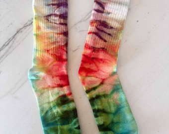 May include: A pair of ribbed crew socks featuring a tie-dye pattern with blended colors of purple, yellow, orange, red, and green. The socks have a vertical ribbed texture throughout the leg and foot, displaying a vibrant, multi-colored gradient effect.