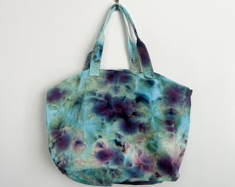 May include: A fabric tote bag featuring a tie-dye pattern in shades of light blue, deep purple, and dark green. The bag has two matching fabric handles and a wide, rounded body shape, displaying a soft, mottled color effect across the entire surface.