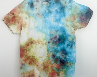 May include: A tie-dye t-shirt in shades of blue, white, brown, and green hangs on a white hanger. The shirt's design features a blend of colors, creating a unique, artistic pattern. The t-shirt is a casual wear item.
