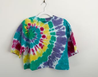 May include: A short-sleeved, tie-dye t-shirt in a spiral pattern. The colors include yellow, green, purple, pink, and teal. The shirt is hanging on a white hanger against a white background.