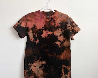 May include: A short-sleeved crew neck t-shirt featuring a tie-dye pattern in shades of black, rust orange, and muted pink. The garment is displayed on a white plastic hanger, showcasing a mottled, irregular color distribution across the fabric.