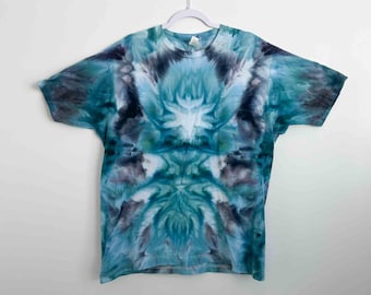 May include: A short-sleeved t-shirt with a tie-dye pattern in shades of blue, teal, and gray. The shirt is hanging on a white hanger against a white background. The tie-dye design is symmetrical, with a central floral-like motif.